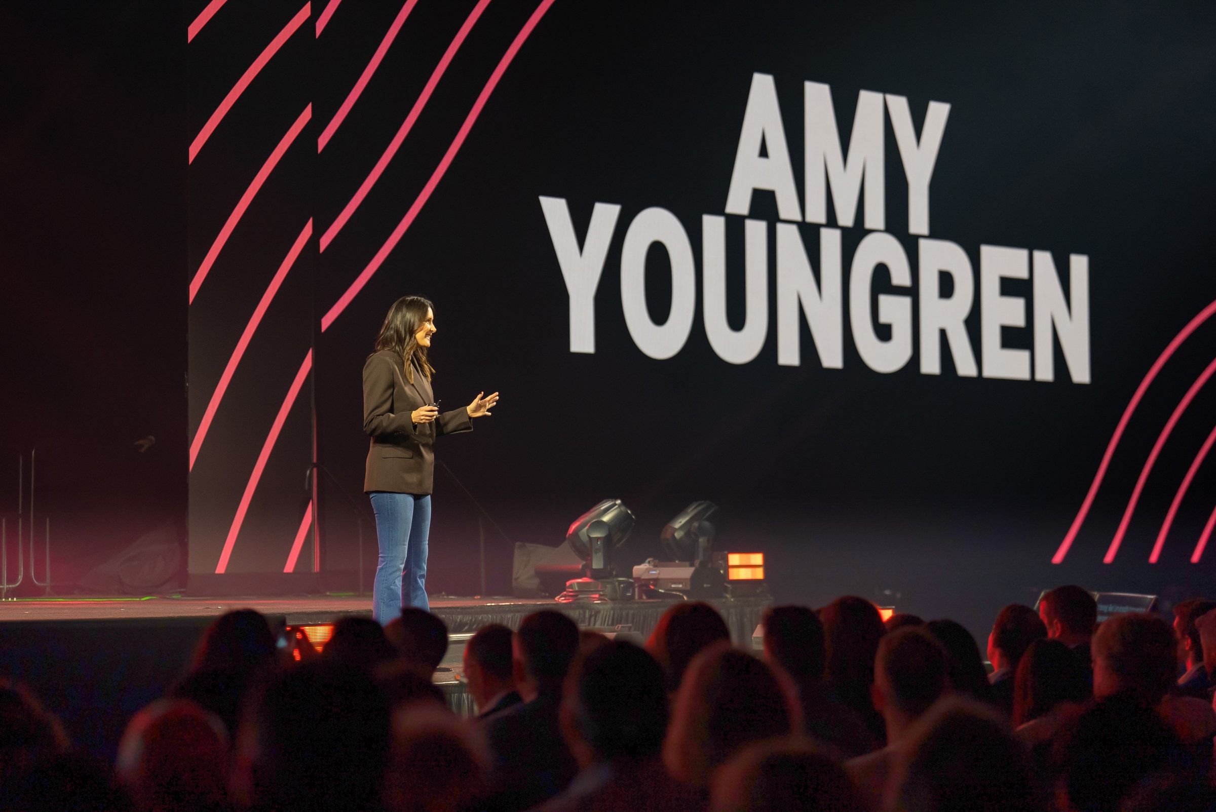 Amy Youngren speaking on a large stage to a full room, her name on the screen behind her